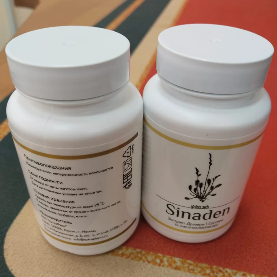 Две закрытые баночки с капсулами Sinaden стоят на столе: одна повёрнута лицевой стороной, другая - обратной с информацией о производителе, условиях хранения, противопоказаниях и т.д.