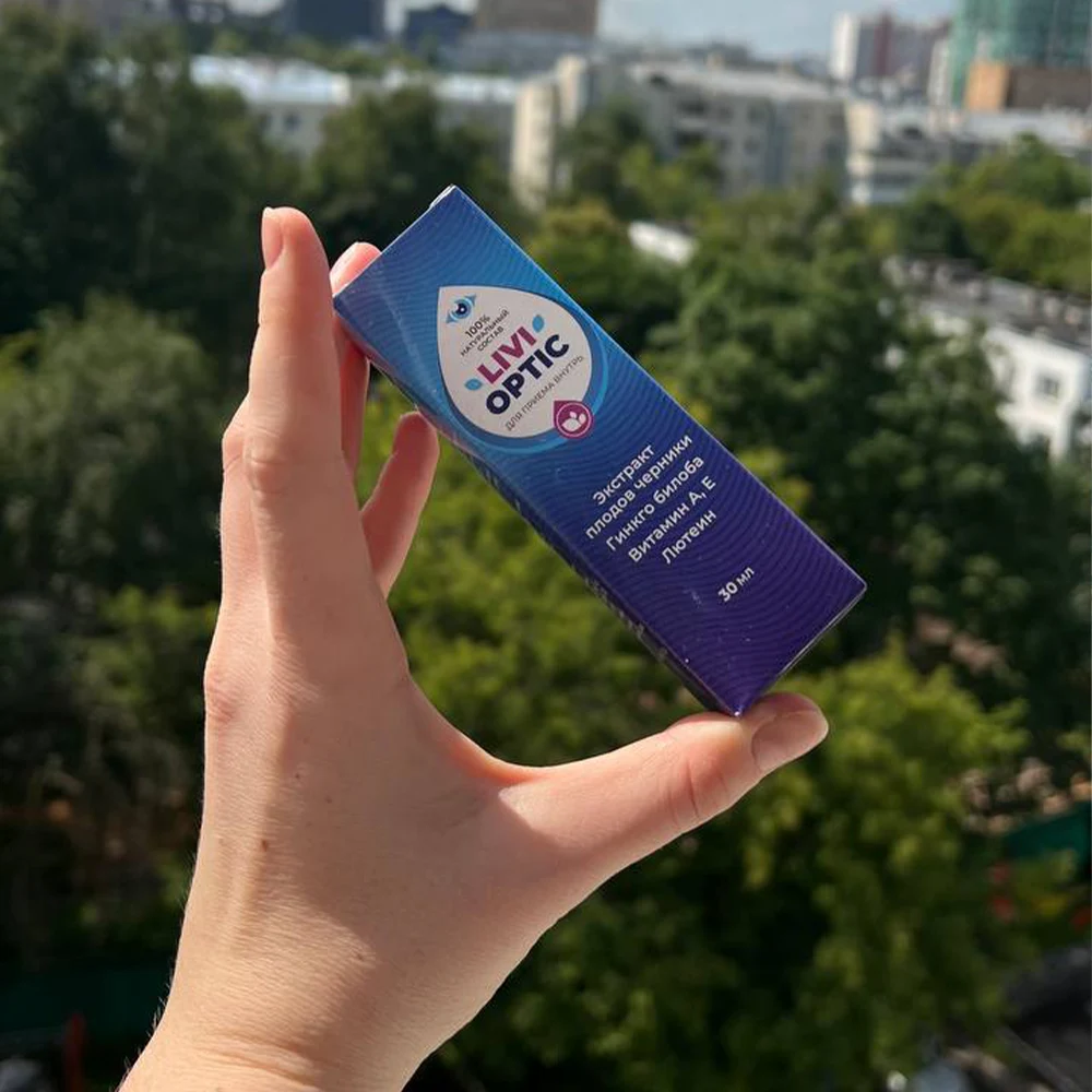 Рука держит упаковка с каплями Livioptic на фоне городских деревьев