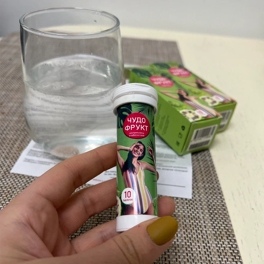 Женская рука держит тубу с таблетками Чудо фрукт на фоне стоит стакан с водой, лежит инструкция и две упаковки Чудо фрукта