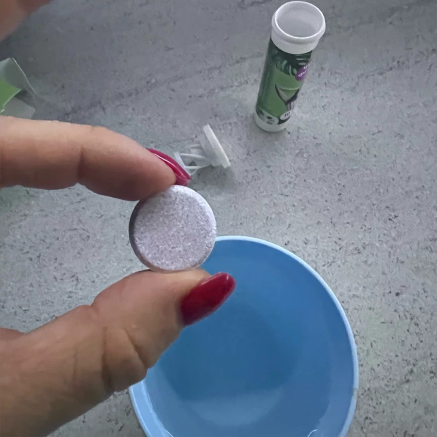 Женская рука держит растворимую таблетку Чудо фрукт на фоне голубой кружки с водой и открытой тубы с шипучими таблетками Чудо фрукт