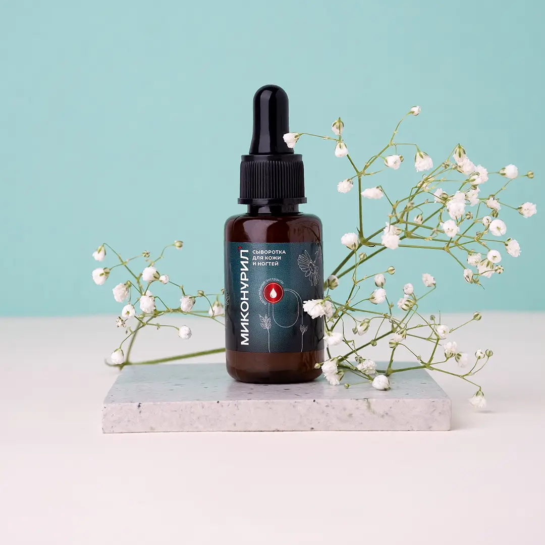 Темно-коричневый флакон с пипеткой и надписью 'МИКОНУРИЛ' на бирюзовой этикетке, содержащий сыворотку для кожи и ногтей. Флакон стоит на мраморной подставке в окружении нежных белых цветов (гипсофила). Фон - мятно-бирюзовый.