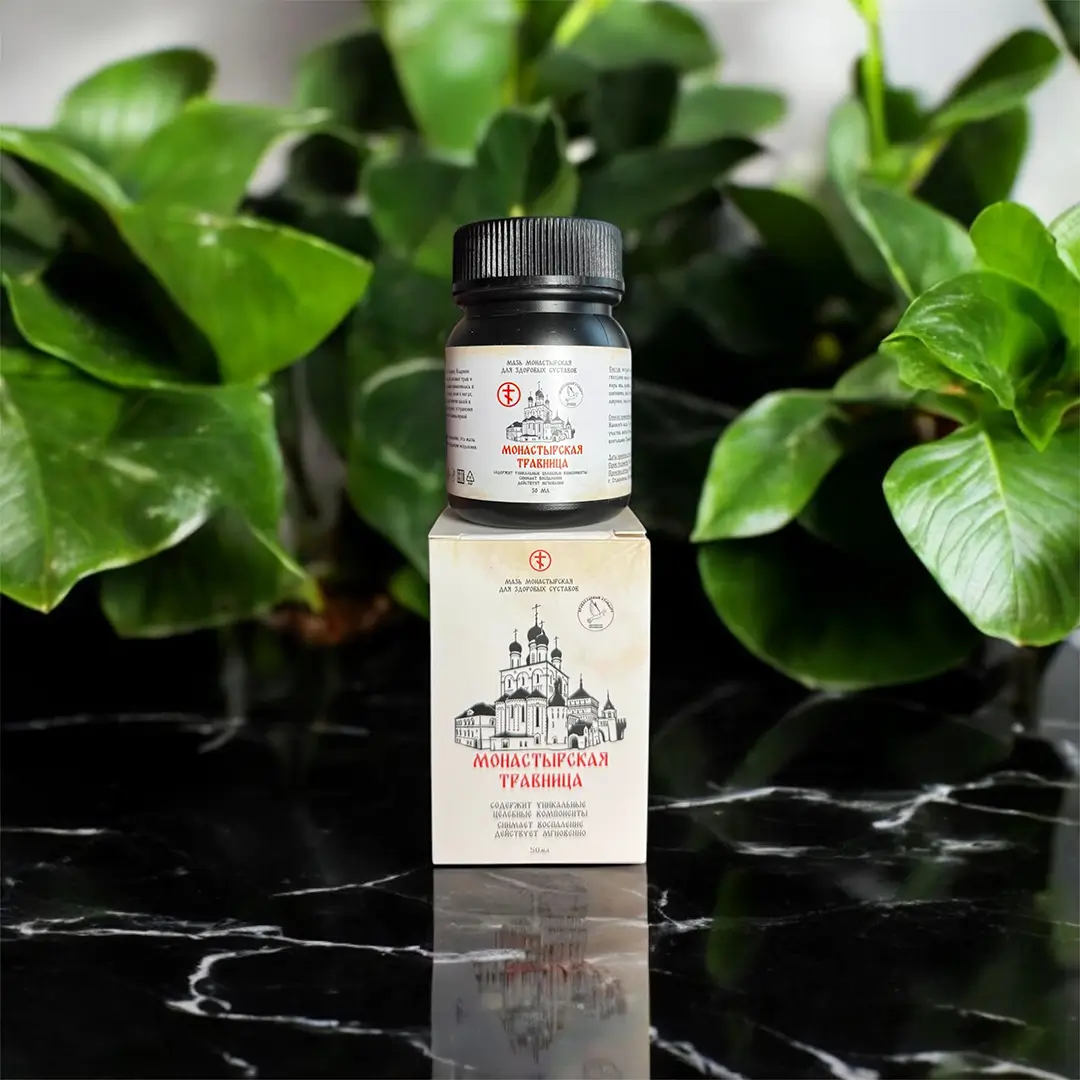 Банка мази Монастырская травница стоит на упаковке среди зеленых листьев растений на мраморной поверхности